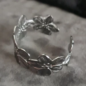 Blommig silverarmband justerbar - Snyggt armbandi silverfärgad metall med detaljerade blommor runt hela bandet. Den har ett öppet band som gör att storleken kan justeras. Perfekt för dig som gillar unika och naturinspirerade smycken med lite extra attityd.