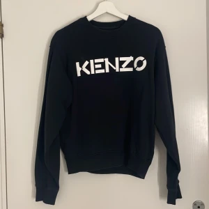 Svart Kenzo sweatshirt med logga - Svart sweatshirt från Kenzo i storlek S med stor vit logga framtill. Klassisk rund halsringning och långa ärmar. Tillverkad i mjuk bomull som är skön att bära. Perfekt för dig som gillar streetstyle och vill ha en clean look.