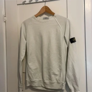 Vit grön sweatshirt från Stone Island Junior - Vitgrön sweatshirt från Stone Island Junior med klassisk rund hals och ribbade muddar. Tröjan har lång ärm och den ikoniska Stone Island-patchen på vänster arm. Tillverkad i mjuk bomull som är skön att bära. Perfekt för dig som gillar clean och stilren streetwear.