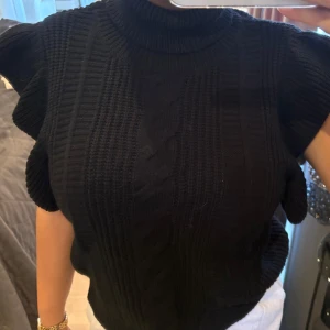 Svart stickad ärmlös tröja - Svart stickad tröja med hög hals och breda ribbade partier. Tröjan är ärmlös med volangliknande axlar och har ett klassiskt kabelstickat mönster framtill. Perfekt för lager-på-lager och snygg till både jeans och kjol.