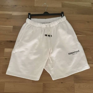 Vita Essentials shorts med snörning - Säljer ett par vita shorts från Essentials Fear of God med elastisk midja och snörning. De har en avslappnad passform och är perfekta för sommaren. Loggan är tryckt på ena benet och materialet känns mjukt och bekvämt.