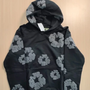 Svart hoodie från Denim Tears x DSM - Svart hoodie från Denim Tears i samarbete med Dover Street Market. Tröjan har stora gråa blommönster över hela och en rymlig huva. Tillverkad i USA i mjuk bomull, perfekt för dig som gillar streetwear med unik design.