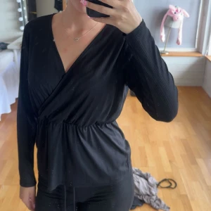 Svart omlottopp med v-ringning - Säljer en svart omlottopp med v-ringning och långa ärmar. Toppen har en markerad midja och är tillverkad i ett mjukt, lätt material som faller snyggt. Perfekt för dig som gillar stilrena och enkla plagg med en twist.