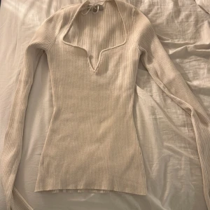 Beige ribbad långärmad topp H&M XS - Supersnygg beige ribbad topp från H&M i storlek XS. Långärmad med v-ringning och knytband i halsen som ger en unik touch. Figurnära passform och mjukt material som sitter skönt. Perfekt att styla med jeans eller kjol för en trendig look.