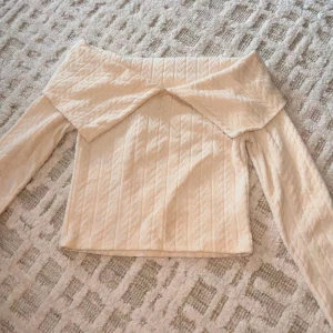 Beige offshoulder stickad topp - Superfin beige offshoulder topp med stickat mönster och bred krage som vilar över axlarna. Långärmad och croppad modell som ger en trendig vibe. Perfekt att styla med höga jeans eller kjol för en snygg look.