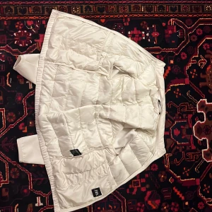 Vit quiltad pufferjacka från Zara - Snygg vit pufferjacka från Zara med quiltad design och långärmade ärmar. Jackan har dragkedja och är tillverkad i ett glansigt syntetmaterial som ger en clean och modern look. Perfekt för dig som vill ha en stilren och varm jacka till vintern.