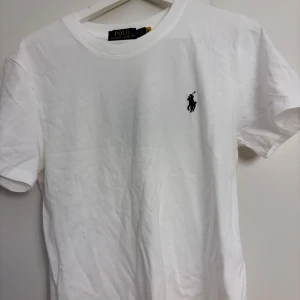 Vit t-shirt från Polo Ralph Lauren - Vit t-shirt från Polo Ralph Lauren med klassisk svart broderad logga på bröstet. Rund hals och korta ärmar, tillverkad i mjuk bomull. Enkel och stilren design som funkar till allt. Snygg passform och skön kvalitet.