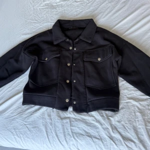 Svart overshirt med tryckknappar - Svart kort overshirt med klassisk krage och stora bröstfickor. Jackan har tryckknappar i silver längs hela framsidan och vid ärmsluten. Materialet är mjukt och känns fleece-liknande, perfekt för lager-på-lager. Enkel och stilren design som passar till allt.
