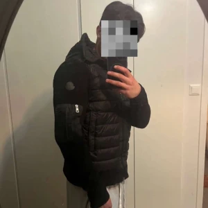 Svart Moncler cardigan med luva  - Säljer en svart pufferjacka från Moncler med stickade ärmar och vadderad framsida. Jackan har huva, dragkedja framtill och en ficka med dragkedja på ärmen. Perfekt för dig som vill ha en stilren och bekväm jacka med snygga detaljer.