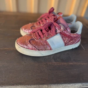 Valentino flycrew  - Snygga sneakers från Valentino Garavani med rosa glitter, breda rosa snören och vita skinnpaneler. Skorna har rund tå, platt sula och små nitar baktill för extra edge. Perfekta för dig som vill sticka ut med en glittrig och cool look.