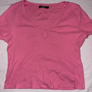 Rosa ribbad topp från Bik Bok - Rosa ribbad topp från Bik Bok med korta ärmar och en söt liten knappdetalj vid nyckelhålsöppningen framtill. Toppen har en croppad passform och är tillverkad i mjukt, stretchigt material som sitter skönt på kroppen.