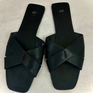 Svarta sandaler - H&M - Stilrena svarta sandaler från H&M i storlek 39. Modell med korsade remmar och fyrkantig tå för en trendig och minimalistisk look. Perfekta till sommaren och passar både till klänning, kjol och jeans.