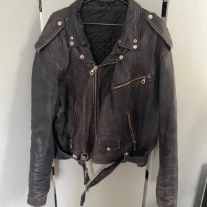 Svart skinnjacka bikerstil XL - Riktigt snygg svart skinnjacka i klassisk bikerstil med dragkedjor, axelklaffar och bälte i midjan. Jackan har flera fickor med dragkedja och quiltat innerfoder. Perfekt för dig som vill ha en edgy look. Sliten till perfektion.