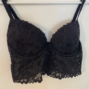 Svart spetsig korsett-topp - Supersnygg svart korsett-topp med blommig spets och vadderade kupor. 