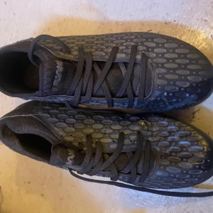 Svarta fotbollsskor från Puma strl 40 - Säljer ett par svarta fotbollsskor från Puma i storlek 40. Skorna har ett coolt prickigt mönster på ovansidan, snörning och rund tå. Yttersulan har fasta dobbar för gräsplan och Puma-loggan syns tydligt undertill. Perfekt för dig som vill ha stil på planen.