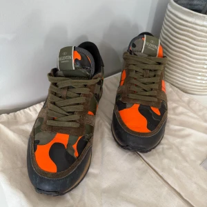 Valentino Rockrunner sneakers camo orange - Snygga Valentino Rockrunner sneakers med orange och grönt kamouflagemönster, svarta och olivgröna detaljer samt mocka- och skinnpaneler. Klassisk snörning och platt sula. Knappt använda där av ett bra pris. Mockan på skorna är helt intakt. 
