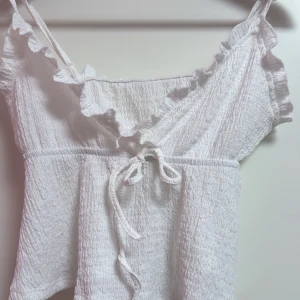 Vit linnetopp med volang och knyt - Supersöt vit linnetopp med volangkant längs urringningen och smala axelband. Toppen har omlottkänsla och knyt framtill, samt en lätt skrynklig struktur i tyget. Perfekt för varma dagar och en romantisk look. Har knappt använt 