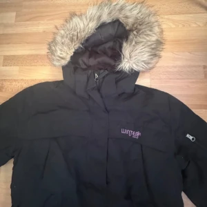 Svart vinterjacka med fuskpäls från Kappahl - Svart vinterjacka från Kappahl Winter Wear med huva och fuskpäls. Jackan har dragkedja och knappar framtill, stora fickor och lång passform. Perfekt för kalla dagar med sin varma och rejäla look. Broderad logga på bröstet och snygga detaljer på ärmen. Det saknas en knapp, men det syns inte. Titta på två sista bilder.