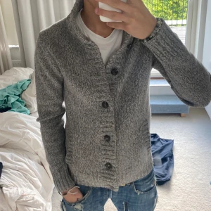 Grå stickad kofta med knappar - Säljer en grå stickad kofta med snygga svarta knappar framtill. Koftan har lång ärm, hög krage och en riktigt chill passform. Perfekt att slänga på sig över en t-shirt för en avslappnad look.