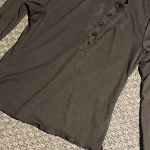 Svart ribbad långärmad tröja med knappar - Svart långärmad tröja med ribbad struktur och flera svarta knappar framtill. Tröjan har en enkel och clean look, perfekt för lager-på-lager. Materialet känns mjukt och stretchigt, vilket gör den skön att bära hela dagen.