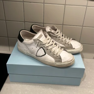 Philippe Model vita sneakers i skinn - Snygga vita sneakers från Philippe Model Paris med grå mockadetaljer och svart häl. Klassisk låg modell med snörning och rund tå. Skorna har en ikonisk sköld på sidan och är tillverkade i skinn och mocka för en lyxig känsla.