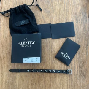 Valentino Garavani svart armband - Snyggt svart armband från Valentino Garavani med coola silverfärgade nitar runt hela bandet. Gjort i skinn/läder med justerbart spänne i silver. Perfekt accessoar för att se skön ut på stranden. Levereras med originalförpackning och dustbag.
