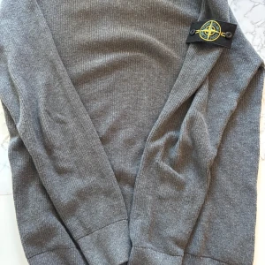 Stone Island stickad sweatshirt  - Säljer en grå stickad tröja från Stone Island i storlek M Tröjan är ribbad och har den klassiska Stone Island-loggan på ärmen. Tillverkad i 100% bomull, perfekt för dig som gillar stilrena och snygga plagg med street-vibe.