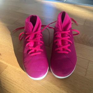 Adidas rosa fotbollsskor med ränder - Säljer ett par snygga Adidas fotbollsskor i stark rosa med svarta ränder och vita sulor. Skorna har snörning och en låg profil som ger bra grepp på planen. Materialet är syntet och designen är riktigt sportig med coola detaljer på hälen. Perfekt för vintervintersäsongen när man spelar inomhus