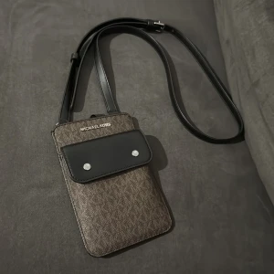 Brun och svart axelväska från Michael Kors - Snygg och praktisk axelväska från Michael Kors i brun och svart. Väskan har en justerbar axelrem och en framficka med knappar. Kedjan på baksidan är för att det ska bli lättare att få tag på allt som ligger i väskan. 