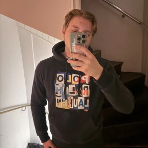 Mörkblå hoodie från Polo Ralph Lauren - Säljer en snygg mörkblå hoodie från Polo Ralph Lauren med färgglad logotyp på framsidan. Tröjan har en klassisk känguruficka och en liten röd logga på ärmen. Perfekt för en avslappnad stil. Sitter mer som S
