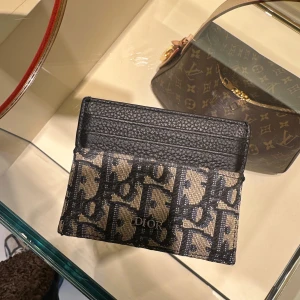 Svart och beige kortfodral från Dior - Säljer ett stilrent kortfodral från Dior i svart och beige med ett ikoniskt mönster. Perfekt för att hålla dina kort organiserade med stil. Fodralet har flera fack och är tillverkat i ett slitstarkt material.