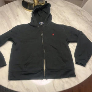 Svart hoodie från Polo Ralph Lauren - Säljer en klassisk svart hoodie från Polo Ralph Lauren med dragkedja och det ikoniska röda logotypen på bröstet. Perfekt för en avslappnad stil. Hoodien har långa ärmar och en bekväm passform.  Storlek XL (barn). 18-20