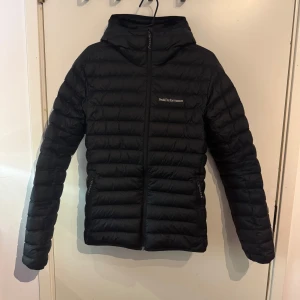 Svart pufferjacka från Peak Performance - Säljer en stilren svart pufferjacka från Peak Performance. Jackan har en praktisk dragkedja och en mysig huva. Perfekt för kyliga dagar med sin quiltade design som håller värmen. Enkelt att matcha med allt i garderoben! Byten är intressant 