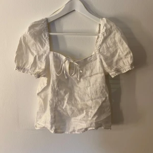 Vit blus med puffärmar - Charmig vit blus med korta puffärmar och en söt knytning framtill. Blusen har en fyrkantig urringning och elastiska detaljer vid ärmslut och rygg för en bekväm passform.