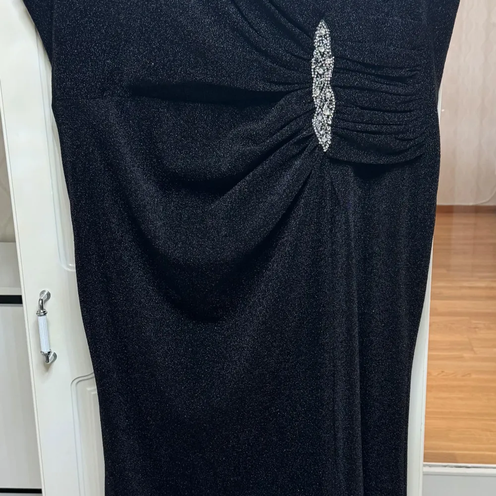 Elegant svart långklänning med glittrande material och en vacker dekorativ detalj framtill. Perfekt för festliga tillfällen. Klänningen har en smickrande passform och är designad för att dra blickarna till sig.. Mekot.