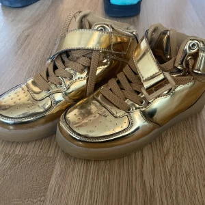 Guldiga sneakers med snörning - Snygga guldiga sneakers med snörning och kardborreband. De har en glansig finish och en bekväm sula. Perfekta för att sticka ut med en unik stil. Den har även blinkande sula