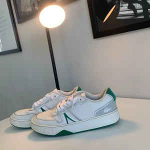 Vita och gröna sneakers från Lacoste - Snygga vita sneakers från Lacoste med gröna detaljer och snörning. Skorna har en klassisk design med perforeringar på ovansidan och en grön sula. Perfekta för en sportig look.