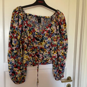 Blommig blus  - Säljer en färgglad blommig blus från Lindex  med långa ärmar och knytband framtill. Perfekt för att liva upp garderoben med sina livliga färger och mönster. Blusen har en lös passform och är tillverkad i ett lätt material.