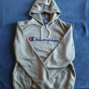 Olivgrön hoodie från Champion - Säljer en olivgrön hoodie från Champion med det klassiska logotypen broderad på bröstet. Tröjan har en justerbar huva med vita snören och en praktisk magficka. Perfekt för en avslappnad stil.