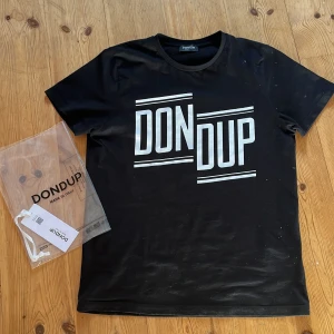Svart t-shirt från Dondup - Säljer en svart t-shirt från Dondup med stort vitt tryck på framsidan. T-shirten har en klassisk rund hals och korta ärmar. Perfekt för en stilren och avslappnad look.Nästan aldrig använd och har inga defekter och är storlek s. Va inte rädd att fråga svarar på allt! 
