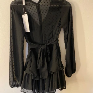 Svart byxdress ifrån Amisu - Elegant svart byxdress från Amisu med  volangdetaljer. Byxdressen har långa ärmar och en v-ringad design. Den är försedd med ett knytband i midjan för en smickrande passform. Det är en byxdress men ser ut som en klänning