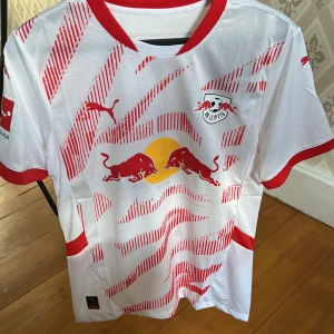RB Leipzig hemma fotbollströja Xavi simons 10 - Säljer en snygg vit och röd RB Leipzig fotbollströja från Puma. Tröjan har korta ärmar och ett coolt mönster med röda detaljer. Den har RB Leipzigs logga och Bundesliga-tryck på armen. Tröjan medföljer i en puma påse