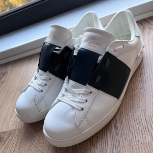 Valentino Open sneakers i vitt och svart - Snygga Valentino Open sneakers i vitt med en bred svart rem över snörningen. Skorna har en stilren design med diskreta detaljer och är perfekta för en trendig look.