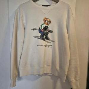 Vit sweatshirt från Ralph Lauren med björntryck - Säljer en vit sweatshirt från Ralph Lauren med ett coolt tryck av en björn som åker skidor. Tröjan har långa ärmar och en rund halsringning. Perfekt för en avslappnad stil eller för att ge en sportig touch till din outfit.
