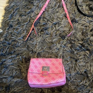 Rosa axelväska från Guess - Säljer en snygg rosa axelväska från Guess med ett mönster av logotyper. Väskan har en justerbar axelrem och en kedjedetalj. Den stängs med en magnetknapp och har flera fack inuti för enkel organisering. Perfekt för att ge din outfit en färgklick! 💖
