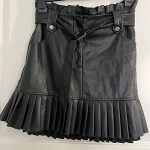 Svart kjol från Zara - Snygg svart kjol från Zara i skinnimitation med plisserad nederkant och bälte i midjan. Kjolen har en cool design med knappar och fickor framtill. Perfekt för att ge din outfit en edgy touch!