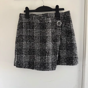 Svartvit rutig kjol med dragkedja - Snygg svartvit rutig kjol med en stilren dragkedja bak och en dekorativ ringdetalj framtill. (Katt o kanin finns i hemmet)