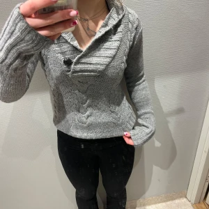 Grå stickad tröja med knappar - Säljer en mysig grå stickad tröja med kabelstickat mönster och stora knappar. Tröjan har en unik krage och långa ärmar, perfekt för kyliga dagar. Passar bra till både jeans och leggings.