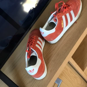 Röda Adidas Gazelle sneakers - Snygga röda Adidas Gazelle sneakers med vita detaljer och snörning. Skorna har den klassiska tre-randiga designen på sidorna och en vit sula. Perfekta för en sportig och stilren look.