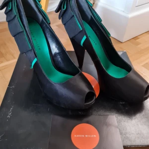 Svarta pumps från Karen Millen - Eleganta svarta pumps från Karen Millen med öppen tå och unik volangdetalj på baksidan. Skorna har en stilren design med en kontrasterande grön insida som ger en modern touch.